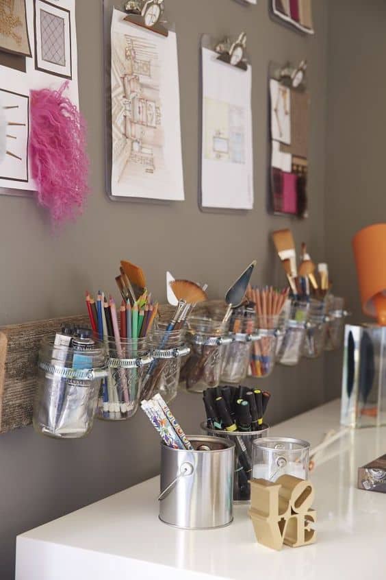 Wall-mounted clear glass jars on a wooden plank display art supplies, enhancing organization and aesthetics in a small office space.