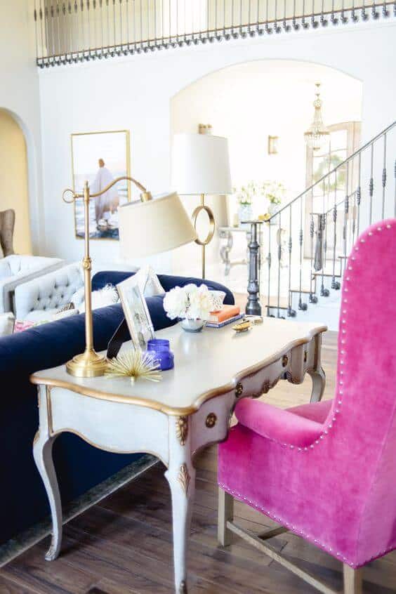 Stylish home office with a distressed white desk, pink armchair, and navy sofa. Bright, elegant design with natural light from arched doorways.