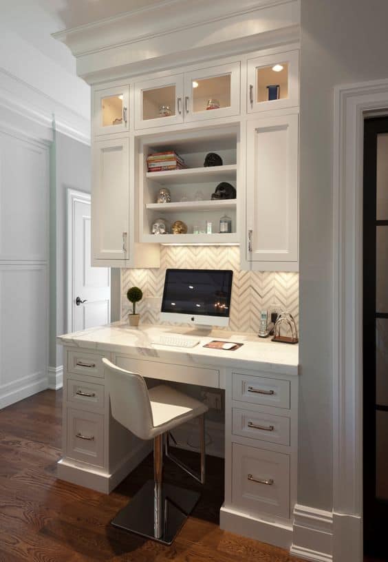 Home office design with a built-in desk, white cabinetry, and shelving for storage. Features a modern chair and warm hardwood flooring.