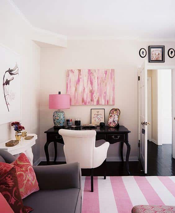 Modern living room office combo with a sleek black desk, vibrant pink abstract art, plush white chair, and gray sofa on a striped rug.