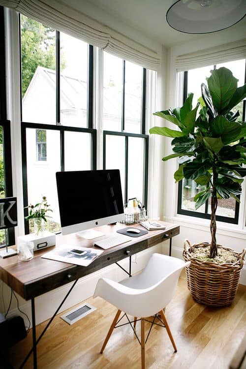 Home office with a wooden desk under large windows, featuring a computer setup, ergonomic chair, and indoor plant, enhancing productivity in a cozy space.