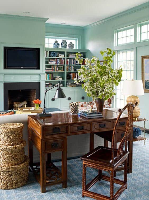 Home office features a wooden desk with intricate carvings against a light teal wall, cozy sofa, and large windows for natural light.