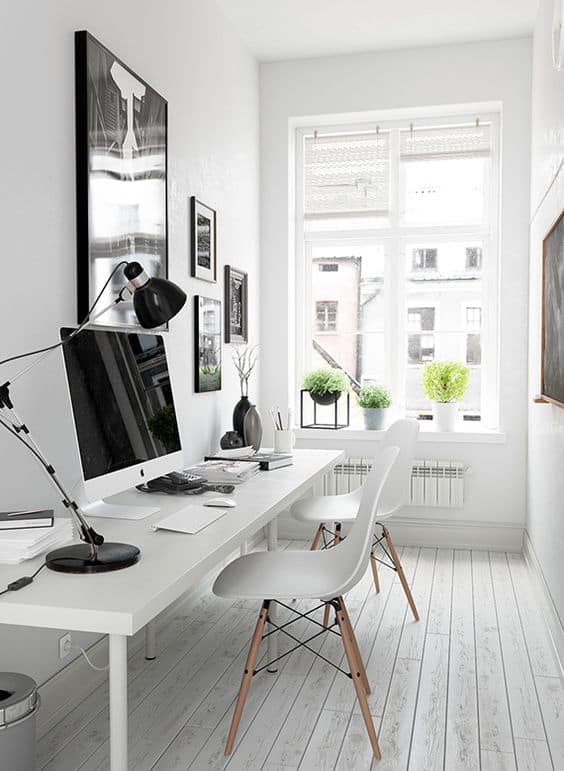 Minimalist small office space featuring a sleek white desk, Apple computer, and modern lamp. Light wood floors and green plants enhance the airy decor.