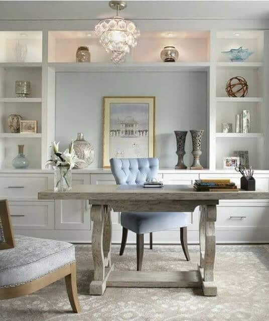 Rustic wooden desk paired with a light blue chair in a tranquil living room office combo, featuring white shelving and elegant chandelier.