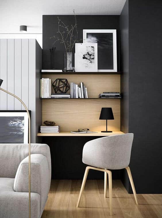 Compact home office decor with a light oak desk, black wall, open shelving, and gray armchair, blending modern style in a living room.