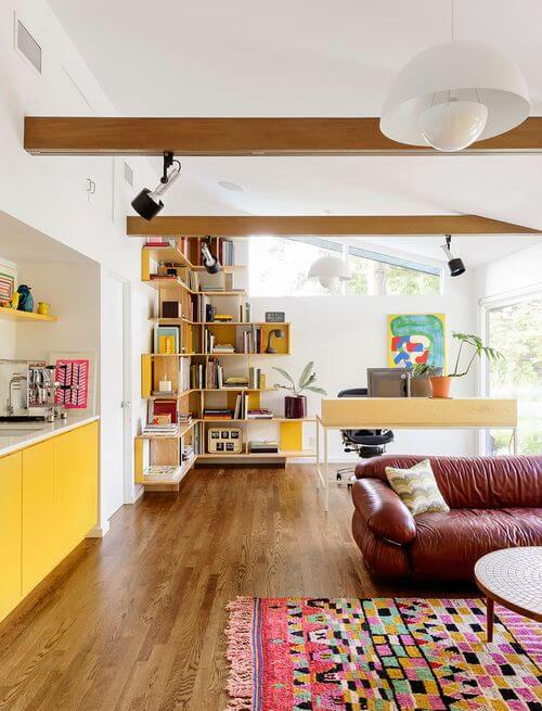 Modern home office featuring a minimalist desk by large windows, asymmetrical bookshelf, and a burgundy leather sofa in a vibrant living room.