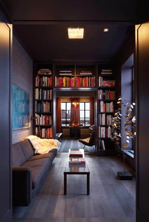 Home library features a dark palette with a plush sofa, coffee table, and colorful bookshelves. Natural light enhances the cozy workspace atmosphere.