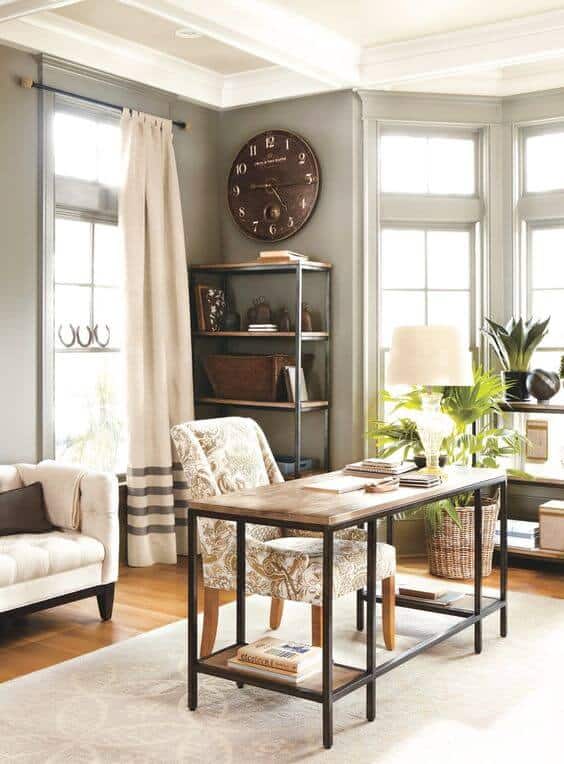 Home office with a reclaimed wood and metal desk, two patterned chairs, a wall clock, metal bookshelf, and a potted plant, filled with natural light.