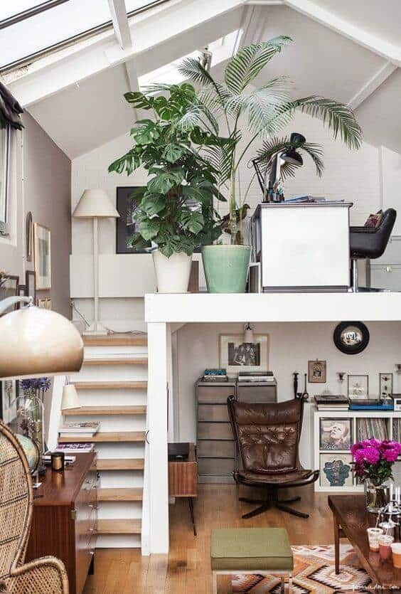 Bright living room office combo with large windows, minimalist desk, tropical plant, cozy rug, and framed artwork, creating a functional workspace.