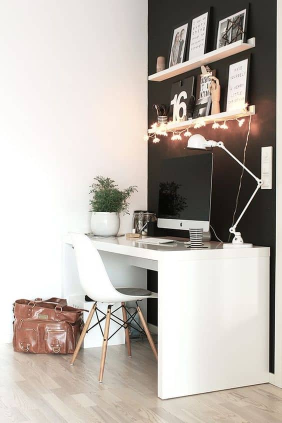 Compact home office with a white desk, minimalist chair, and black accent wall. Features framed art, a desk lamp, and a potted plant for greenery.