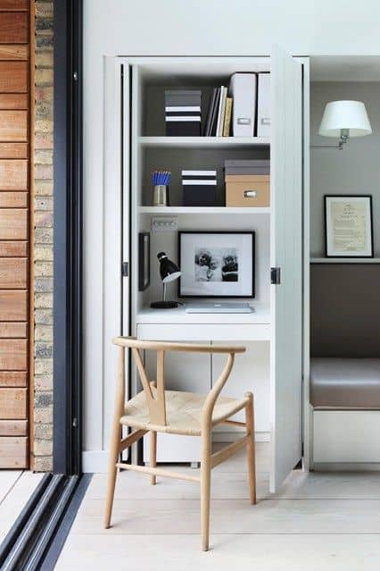 Compact home office nook featuring light wood cabinetry for storage, a white desk with a stylish lamp, and a woven-seat chair by a large window for natural light.