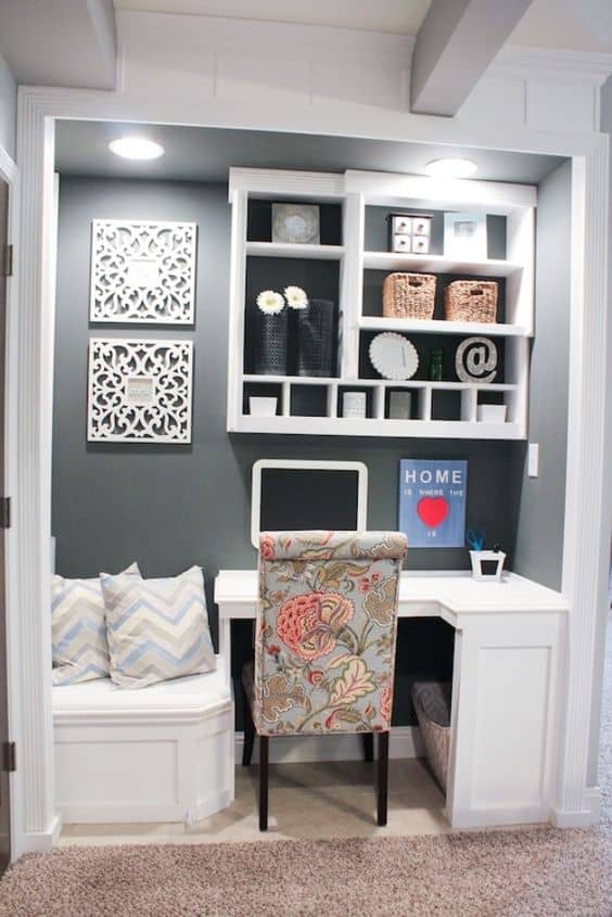 Compact workspace with a white wall-mounted desk against a dark gray backdrop, open shelving, and floral-upholstered chair, enhancing productivity.