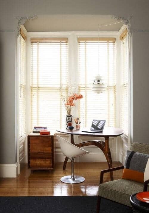 Cozy home office nook with natural light from bay windows, featuring a polished wooden round table, modern white chair, and vintage cabinet.