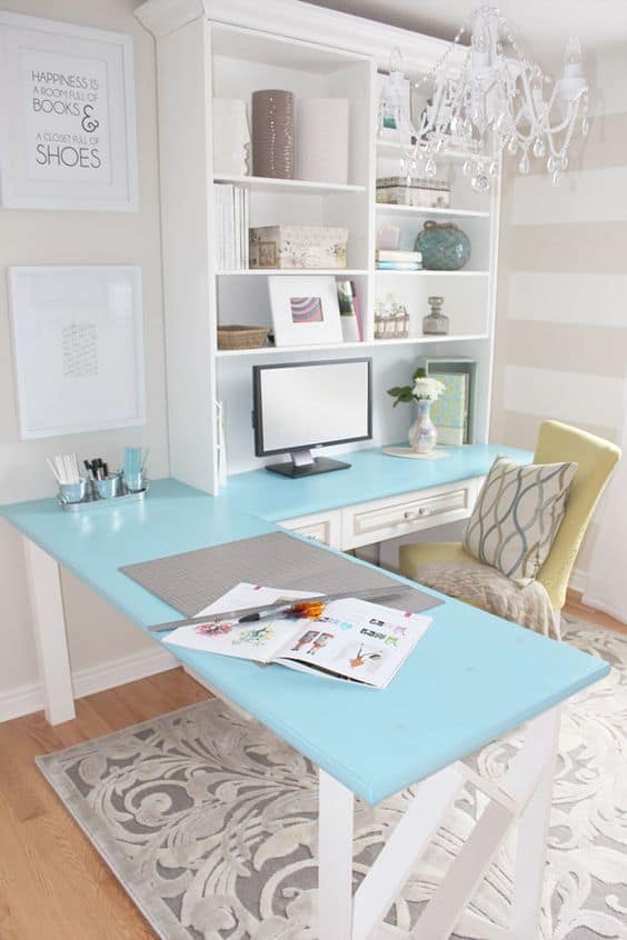 Home office with L-shaped blue desk, white legs, organized stationery, built-in shelving, green accent chair, and beige striped walls.