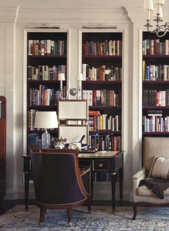 Classic wooden desk with built-in bookshelves in a warm home office. Leather chair faces the desk, with a patterned armchair nearby.