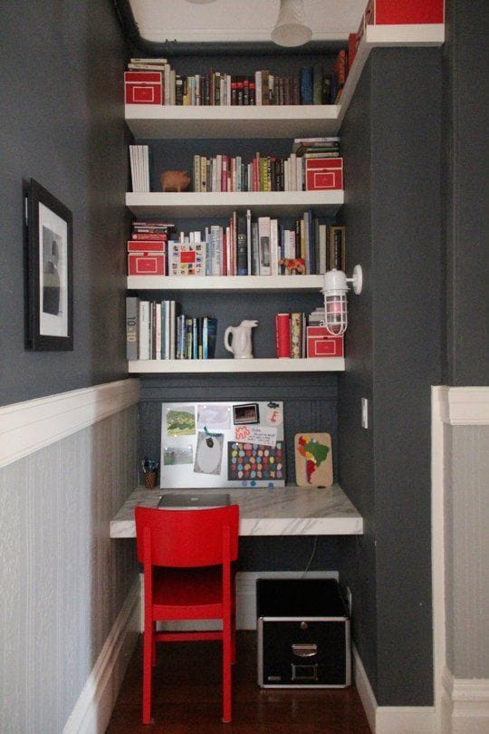 Compact home office features a marble-topped desk, vibrant red chair, and floating shelves for storage, set against a deep gray wall.