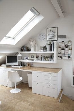 Sleek home office with white cabinetry and natural wood surfaces, featuring a large skylight, integrated desk storage, and open shelving.