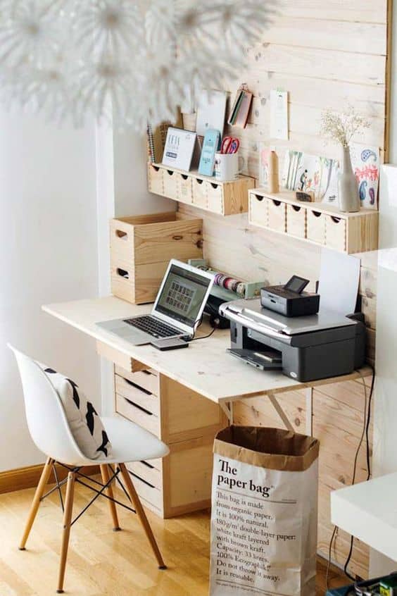 Compact home office with a light wood desk, white ergonomic chair, wooden storage boxes, laptop, and printer, enhanced by natural light.