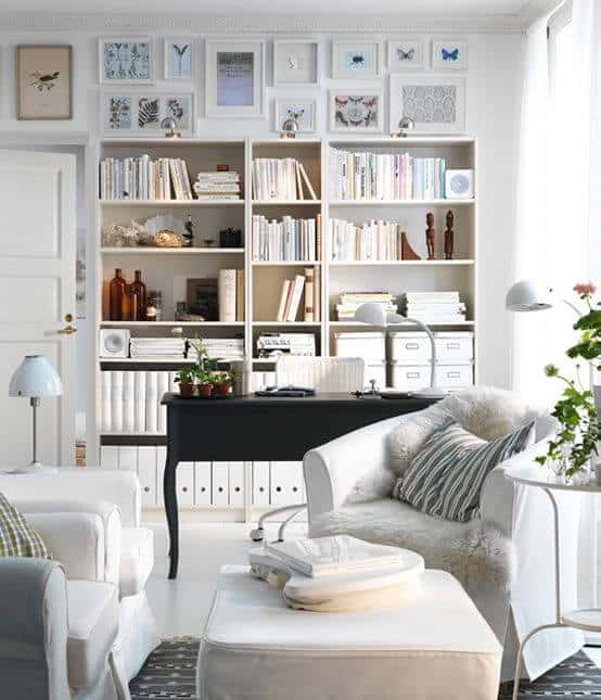 Well-organized living room office combo features a sleek black desk, natural light, light wood bookshelf, and cozy white sofa with pillows.