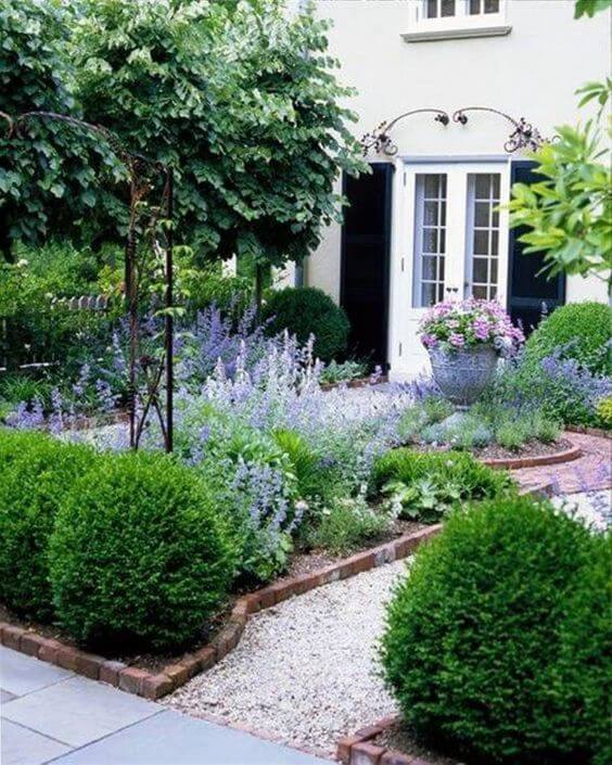 Symmetrical small front garden with lush greenery, rounded boxwoods, lilac blooms, and brick-edged gravel paths, enhancing curb appeal.