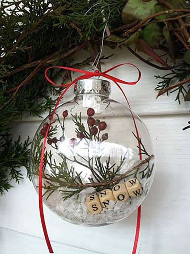 37 DIY Christmas Ornament Ideas For You Unique Decor 11 Clear glass Christmas ornament filled with green sprigs, red berries, and faux snow, hung with a red ribbon for festive decor.