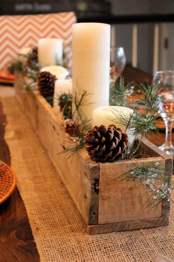 Rustic wooden box centerpiece with cream-colored candles, pinecones, and evergreen sprigs, set on a burlap runner over a dark wooden table.