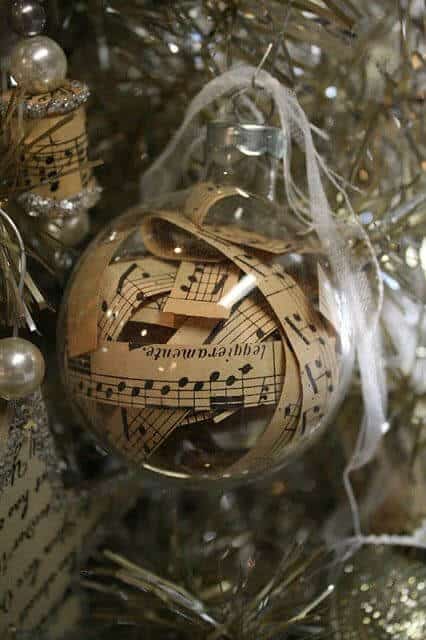 37 DIY Christmas Ornament Ideas For You Unique Decor 21 Christmas ornament with layered sheet music in a glass sphere, tied with a silver ribbon, set against shimmering silver tinsel.