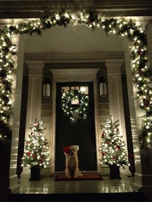 Christmas decorations adorn a welcoming porch, featuring a dark green door, potted evergreen trees with lights, and a festive wreath.
