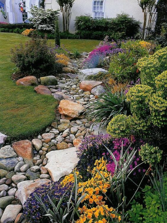 Gently sloping pathway with smooth river rocks, flanked by vibrant yellow, purple, and green flowers, set in lush grass.