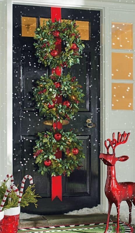 Three stacked holiday wreaths with red ornaments and ribbons adorn a black door, complemented by a red reindeer figurine and falling snow.