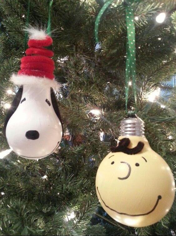 37 DIY Christmas Ornament Ideas For You Unique Decor 29 Snoopy and Charlie Brown ornaments, made of painted glass, hang on a lush green Christmas tree adorned with warm white lights.