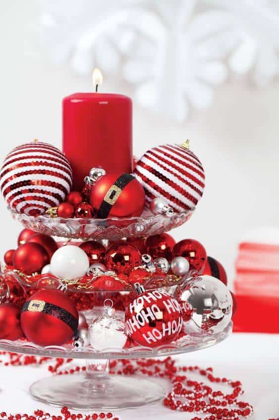 Tiered glass stand adorned with red and silver Christmas ornaments, featuring a bright red candle and decorative bead strands for a festive touch.