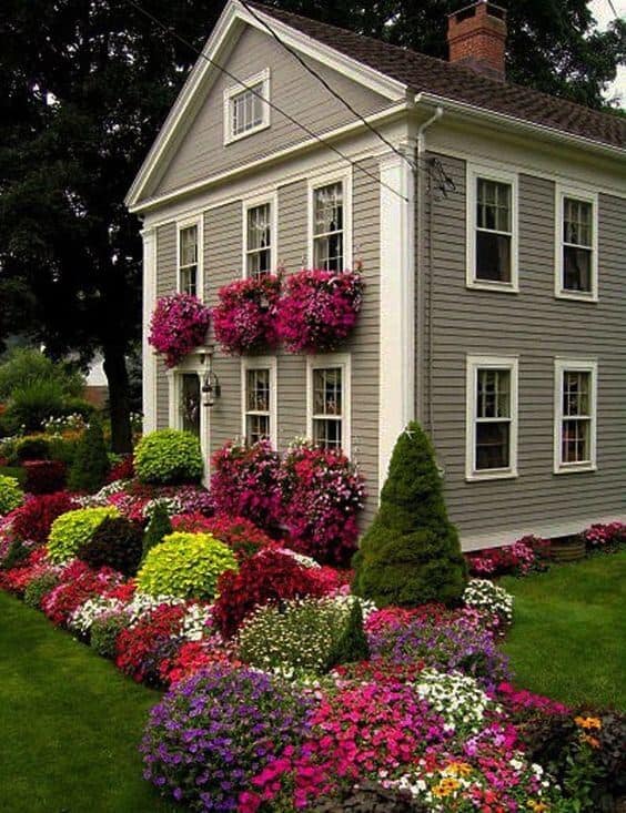 Vibrant flower beds of petunias and begonias surround a charming two-story house, with boxwood hedges enhancing curb appeal.