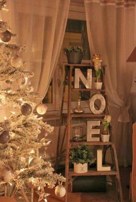 A white artificial tree decorated with silver and gold Christmas ornaments, alongside a wooden ladder displaying 'NOEL' letters and potted plants.