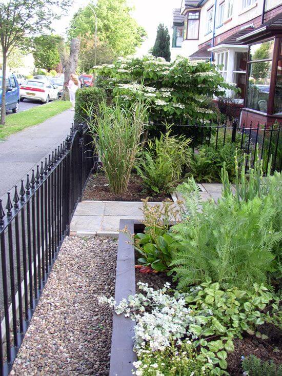 Lush green ferns and colorful flowering plants arranged within a small front garden, bordered by a black wrought iron fence and stone pathways.