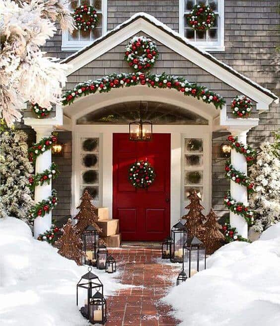 Christmas ornaments decorate a vibrant red door, framed by snow-covered landscaping, with holiday garlands and wooden trees enhancing the festive scene.