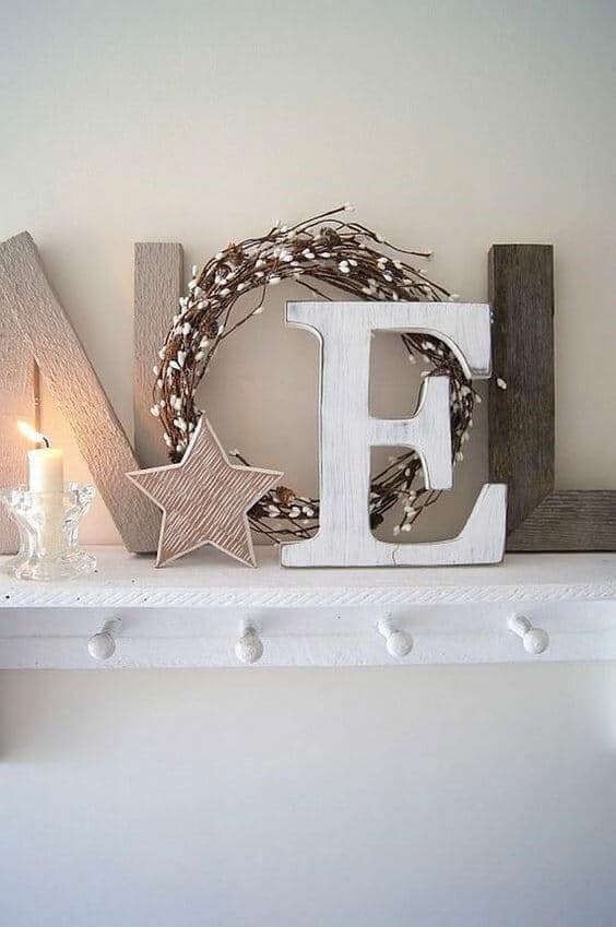 Rustic holiday decor features wooden letters spelling "NOEL" on a whitewashed ledge, with a circular twig wreath and a glowing candle, creating a cozy ambiance.