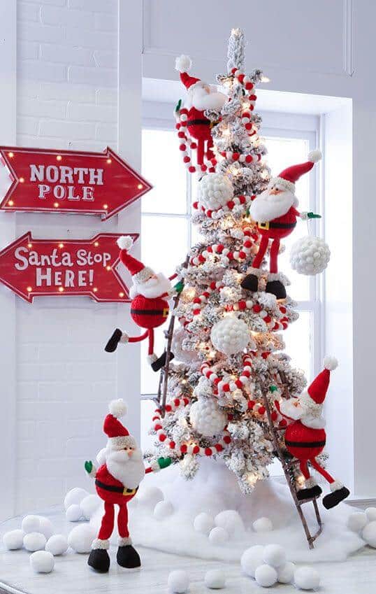 Christmas tree decorated with oversized ornaments and Santa figures, surrounded by faux snow. Cozy indoor setting with festive signs.