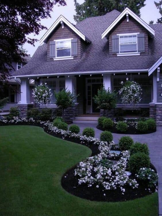 Lush green lawn with manicured shrubs and white blooms in a small front garden, framed by traditional stone and wood house features.