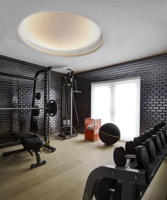 36 Home Gym Ideas - Layouts For Your Perfect Workout Spot 2 Modern home gym featuring strength training equipment, dark brick walls, large windows, and light hardwood flooring for a stylish workout space.