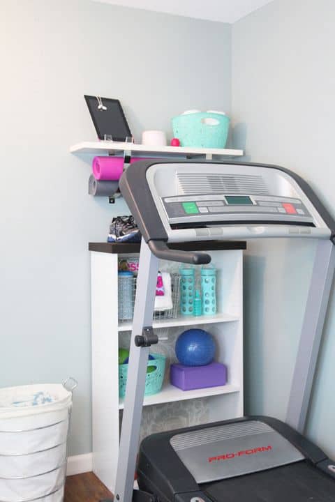 36 Home Gym Ideas - Layouts For Your Perfect Workout Spot 20 Compact home gym with a treadmill against a light gray wall, colorful exercise accessories on a white shelf, and warm wooden flooring.