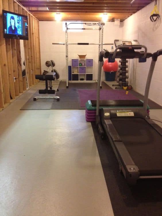 36 Home Gym Ideas - Layouts For Your Perfect Workout Spot 22 Home gym featuring a treadmill and weight bench, rubber flooring, wall-mounted TV, and exposed wooden beams in a bright space.