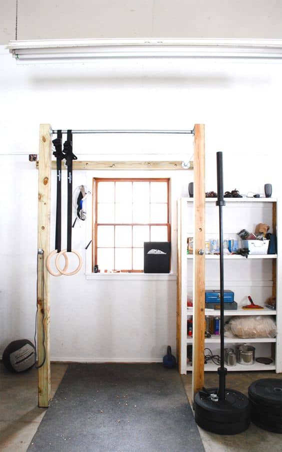 36 Home Gym Ideas - Layouts For Your Perfect Workout Spot 23 Home gym with a wooden pull-up bar and gymnastic rings, rubber flooring, white shelving for weights, and natural light.