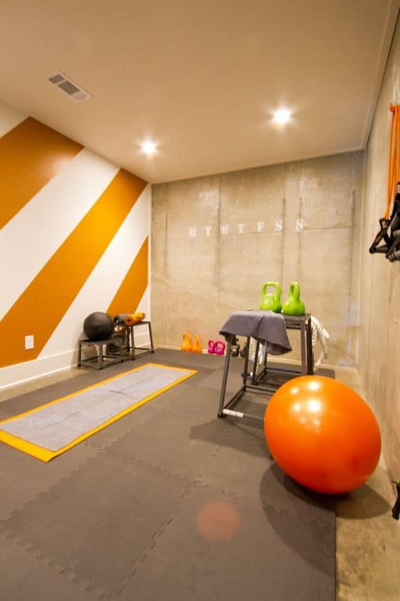 36 Home Gym Ideas - Layouts For Your Perfect Workout Spot 24 Home gym with rubberized flooring, resistance station, weights, orange exercise ball, and yoga mat. Modern design with concrete walls and orange stripes.