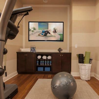 36 Home Gym Ideas - Layouts For Your Perfect Workout Spot 35 Home gym featuring a workout space with a wall-mounted TV, dark wood storage, medium-toned hardwood flooring, and organized fitness accessories.