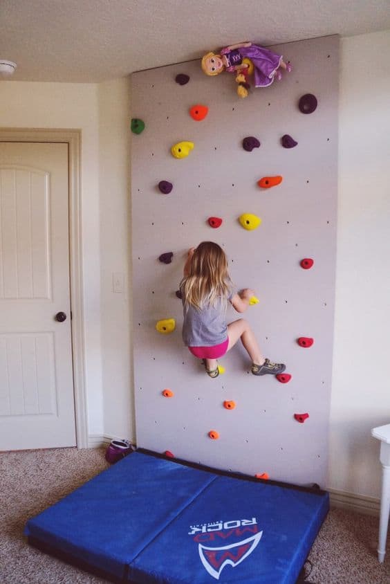 36 Home Gym Ideas - Layouts For Your Perfect Workout Spot 9 Indoor climbing wall with multicolored holds, padded mat below, and cheerful decor in a well-lit room, perfect for kids’ exercise.