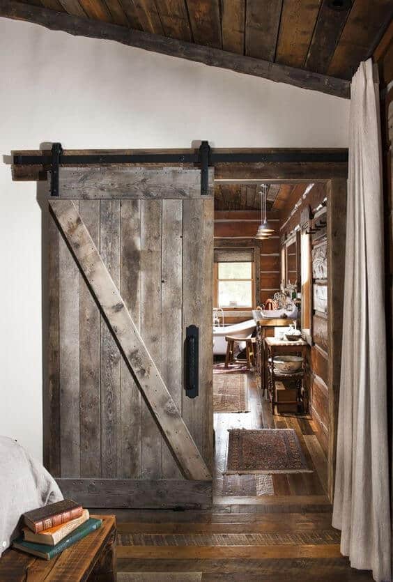Rustic barn door with weathered wood finish, mounted on a black metal rail, enhances cozy farmhouse interior with wooden beams and warm flooring.