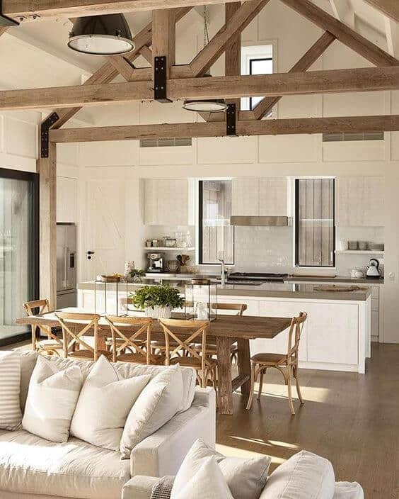 Modern farmhouse kitchen with open concept design, exposed wooden beams, white cabinetry, and a large natural wood dining table. Ample natural light and cozy atmosphere.