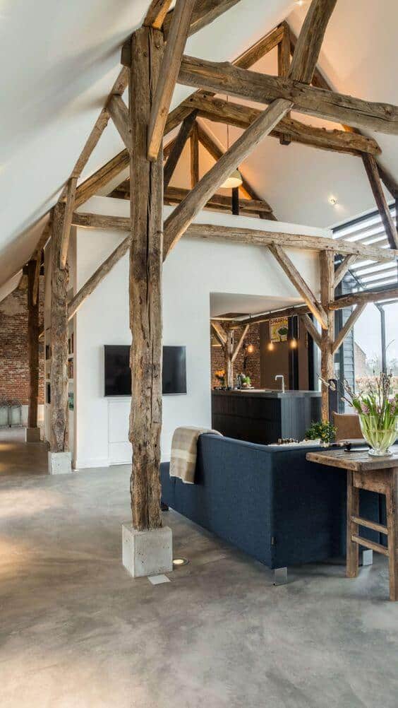Open concept living room with high ceilings, wooden beams, dark blue sofa, reclaimed wood table, and sleek kitchen island. Modern farmhouse design.