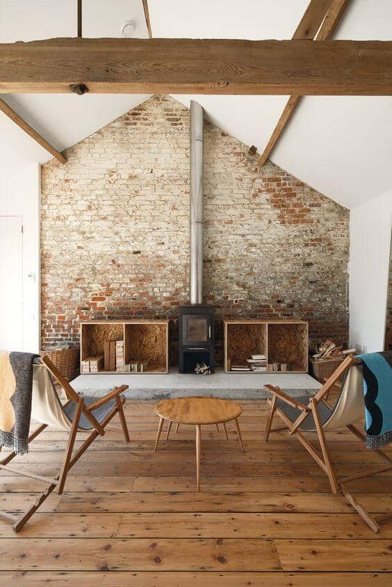Rustic brick wall and exposed wooden beams define this cozy farmhouse interior, featuring folding chairs and a wood-burning stove.